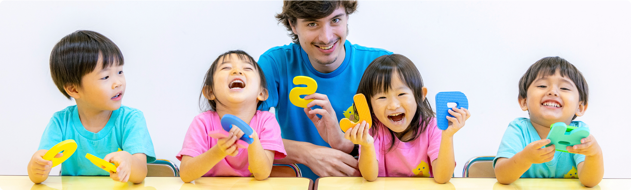 4人の子どもたちがアルファベットのモチーフのおもちゃを持って笑顔で並んで座っている。子どもたちの真ん中には講師が座っている。