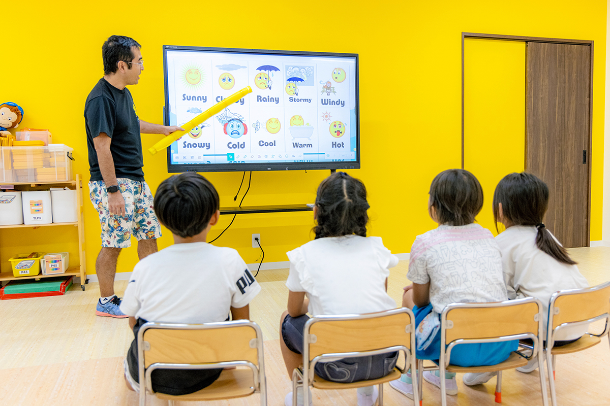 天候を表す英語とイラストがモニターに映し出され、先生が黄色の棒で指し示している写真。4人の子どもたちが先生とモニターの方を向いて椅子に座っている。